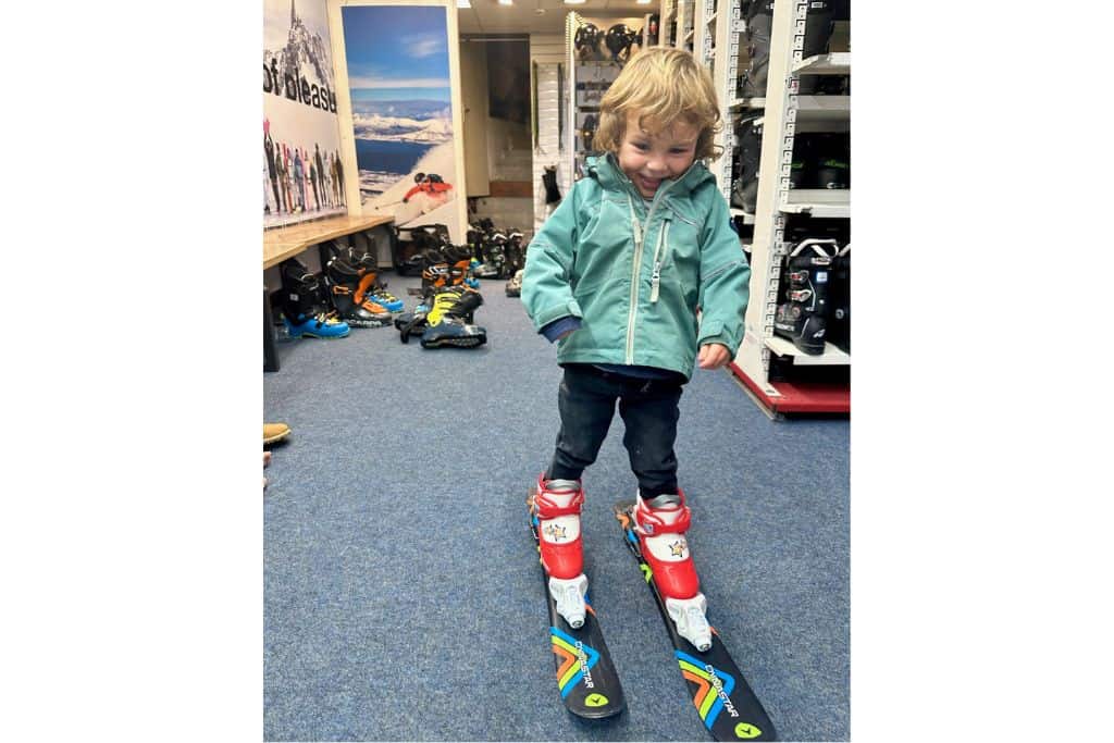 A young boy in a green ski jacket is stood in a ski rental shop trying on his ski boots and skis. He is looking at the camera and smiling. 