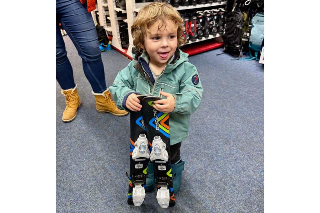 A 3 year old is stood in a ski rental shop with his ski clothes on and looking at the camera as he holds his skiis.