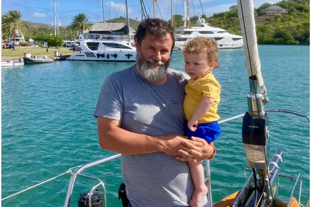 A man is stood on the bow of a sailing yacht wearing a grey t-shirt and in his arms is his  very young son who is wearing a yellow t-shirt and blue shorts. They are both looking at the camera. In the background is Nelson Dockyard and marina with it's buildings.
