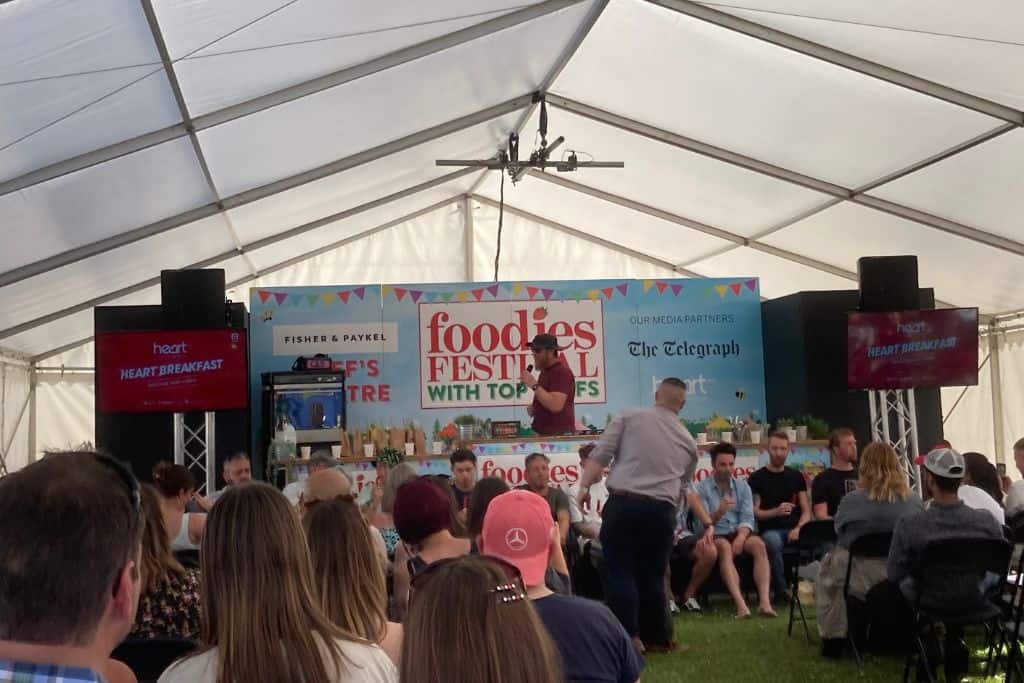 There are lots of people sat in a row in a white marquee watching a small stage where there is a chilli eating competition happening.  In the background is a sign advertising Foodies Festival which is one of the best festivals in Hampshire.