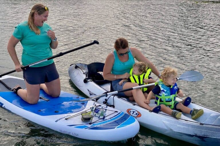 On the left a woman is in a kayak looking down at her son who is sat in front of her. In front of him is another little boy. To the left is another woman laughing who is kneeling on a stand up paddle board. They are kayaking on the River Hamble with kids.