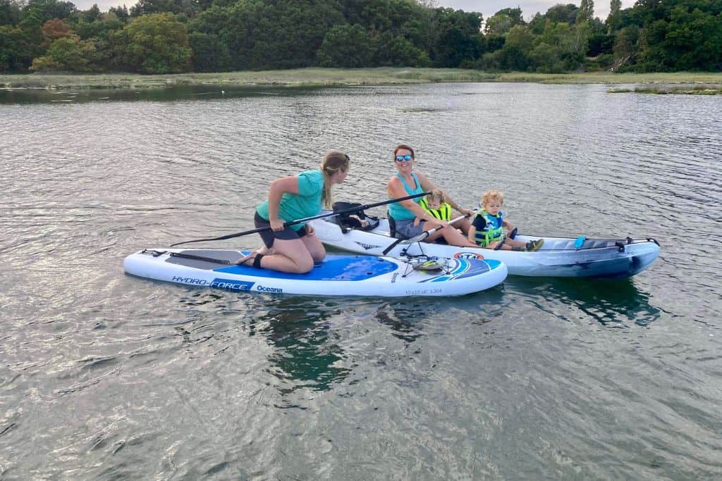 In the front is a woman kneeling on a stand up paddle board facing to the left and chatting to the woman next to her. This woman is in a kayak and smiling at the camera. In front of her are two very young boys.  She is kayaking with the kids on the River Hamble.  In the very background is the shore and some green trees and bushes.