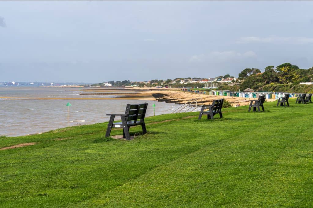 To the right is a large gree grass area that is overlooking the beach and sea to the left.  at the edge are two benches and the background is Lee-On-Solent which is one of the sandy beaches in Hampshire.