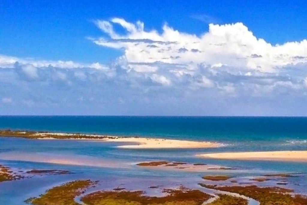 This is an image looking out to sea.  There is a blue sky with some clouds in it. In the water are several sand bars and some marshland. This is the Rio Formosa National Park in the Algarve.