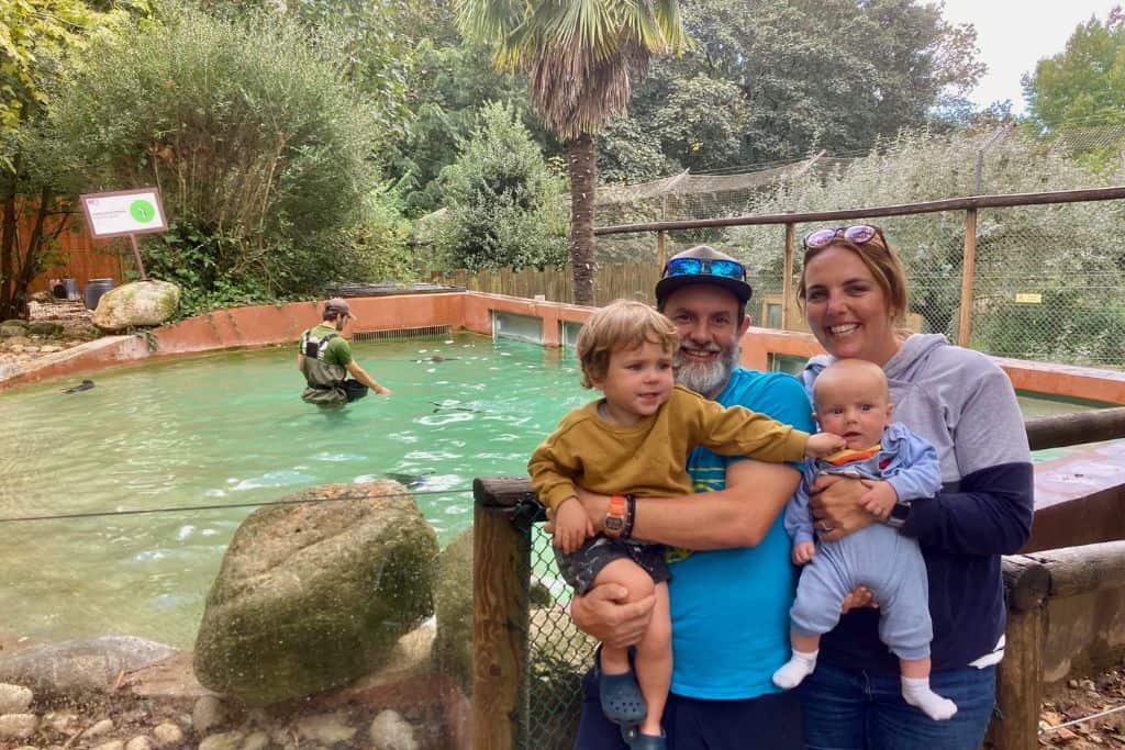 A man and a woman are standing next to each other, he is holding his 3 year old son and she is holding their baby. They are stood in front of a small pool and in the pool are penguins. They are at the Lagos Zoo which is one of the best things to do with Kids in the Algarve.