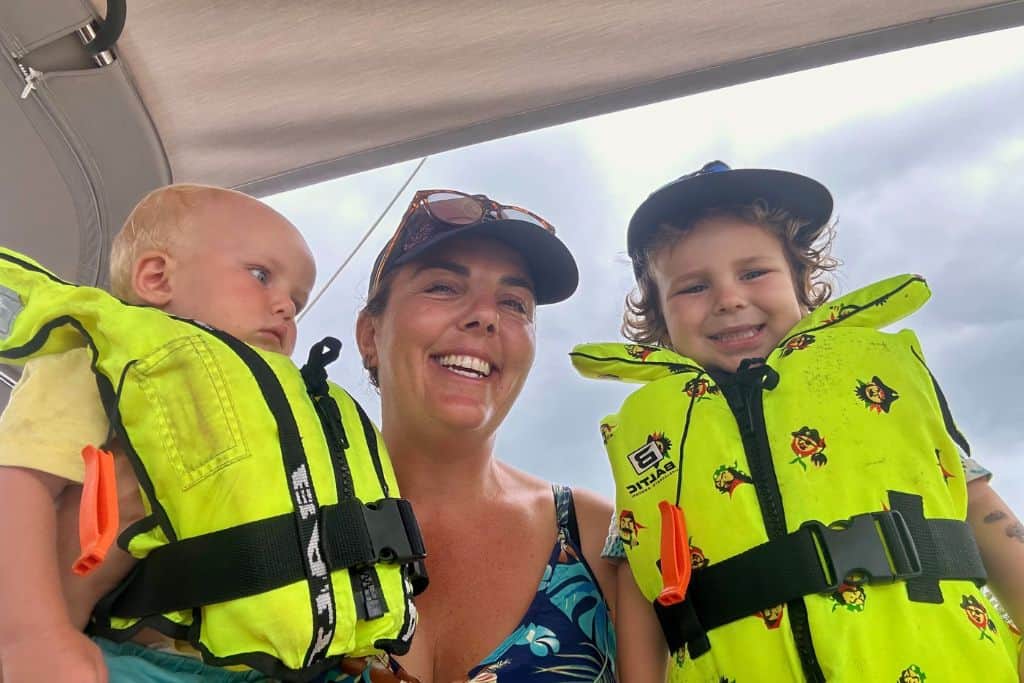 A mum is in the middle of her three year old and baby boys. The boys are wearing bright yellow lifejackets and smiling at the camera. The mum has on a flowery vest top and a baseball cap. She is sailing with her kids.
