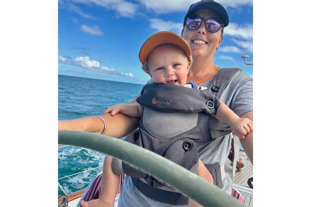 A woman with her baseball cap and sunglasses on with a grey t-shirt is holding the helm of a yacht. In a child carrier is her baby who is smiling at the camera. She is sailing with her baby for the first time.