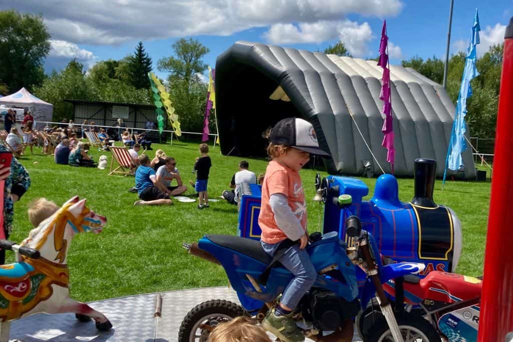 In the front of the image a boy is sat in a motorbike on a mini fairground ride. In the background is a music stage and people are sat there watching a band play.