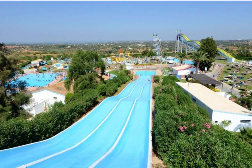 The image is taken looking down a long blue slide at a water park. At the end of the slide is the rest of the water park in the background of the image.  This is Aqualand which is one of the best things to do in the Algarve with kids. 