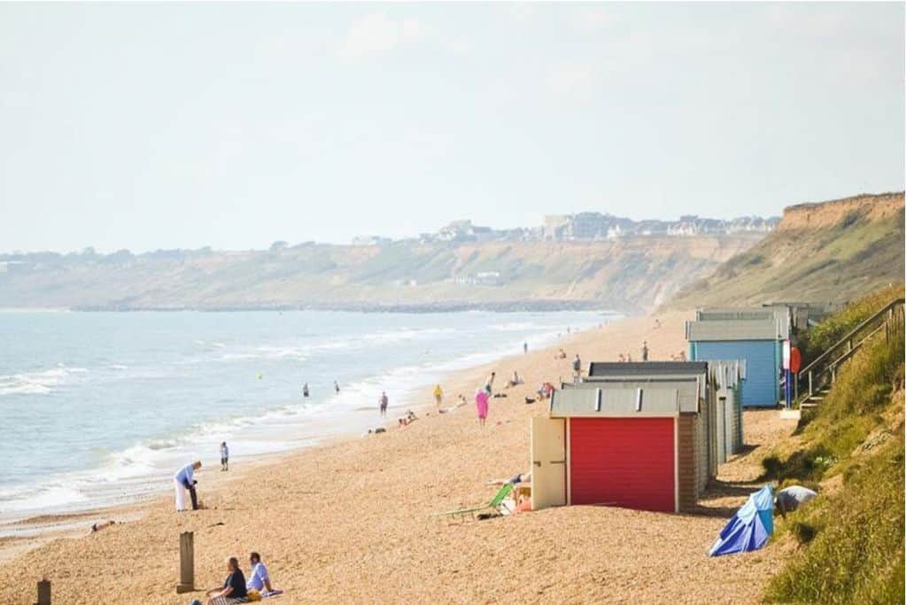 On the left is the sea, and on the right is a sand and shingle beach with some colourful beach huts along it. Behind the huts is a small grassy hill that backs onto the beach. there are people walking along the beach.  This is Barton on Sea which is one of the best sandy beaches in Hampshire.
