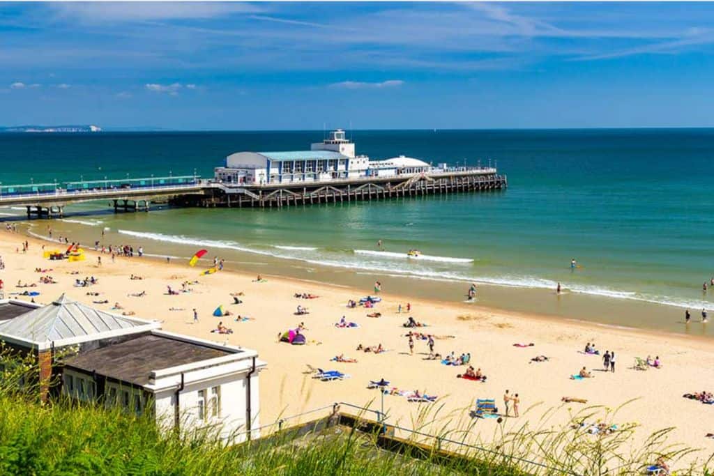 This is taken from above Bournemouth Beach with the beach below with people sunbathing and enjoying the beach. There are also parasols and windbreakers.  On the left going into the sea is Bournemouth Pier with a large white building at the end of the pier.