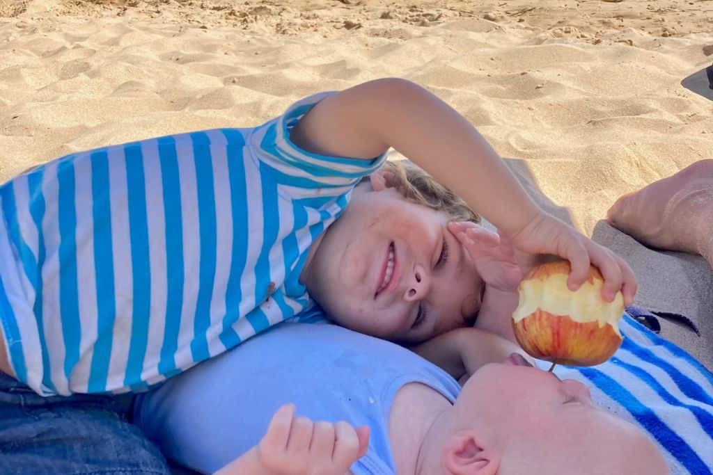 A little boy in a striped t-shirt is lying on his side on a towel on the beach. In front of him, also lying down is a baby and he's dangling a partially eaten apple in the babies mouth.
