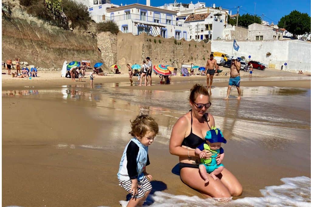 A woman is kneeling in the sea in a black bikini. In her arms is her baby looking at the sea. Next to her is her toddler son who is in his swimwear and laughing as the water comes in. Behind them is old walls and the beach town of Burgau. This is one of the best beaches to go in the Algarve with kids.
