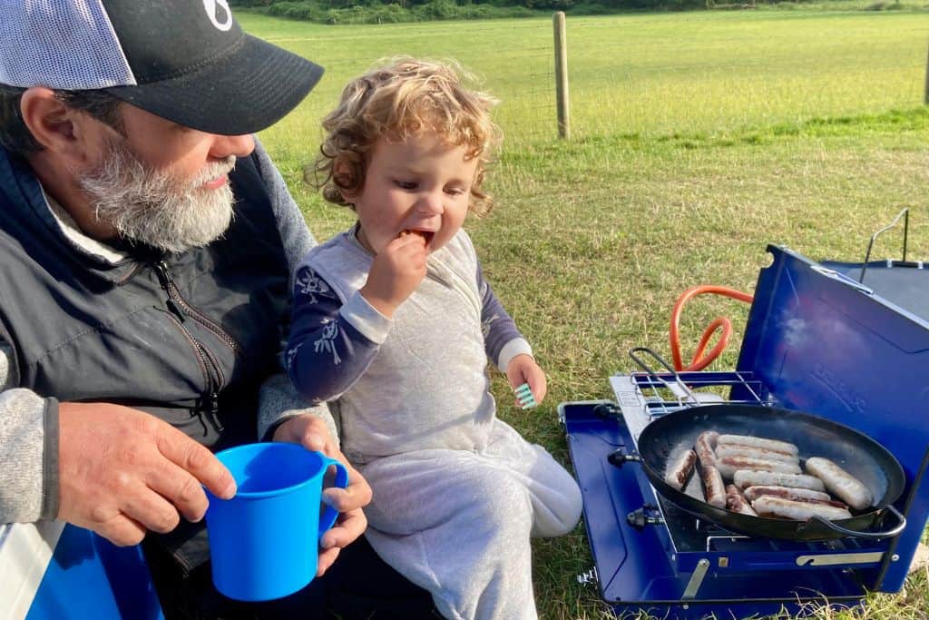 A father is sat on the floor wearing a baseball cap and holding a blue plastic cup. His son is sat on his lap wearing a kids sleeping bag. They are cooking sausages on a frying pan on a gas camping cooker. He is camping with his kids.