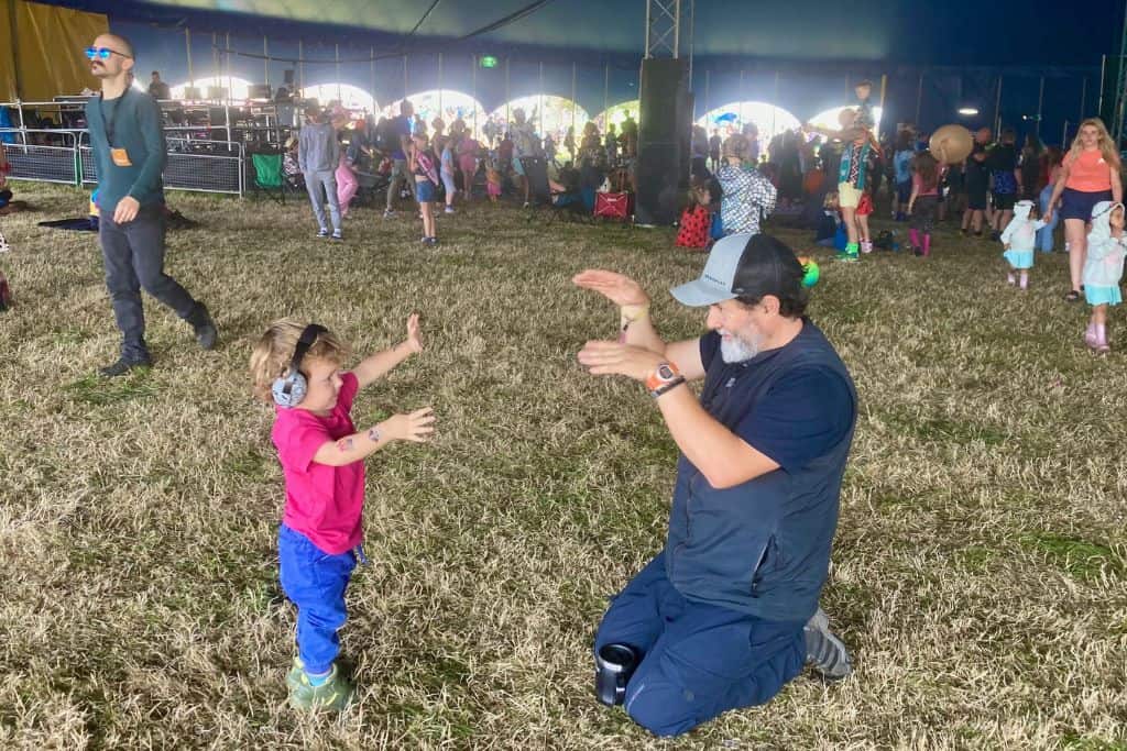 On the left is a boy in a pink t-shirt and blue trousers, he is also wearing ear defenders. Opposite him is a man with a t-shirt, trousers and baseball cap.  They are both dancing to the band that is playing behind them in the marquee that they are in. 