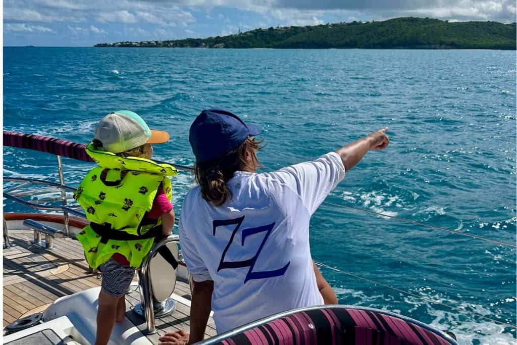 A three year old boy wearing a yellow lifejacket with pirates on it has his back to the viewer and is looking to see from the back of a sailing yacht. Next to him is woman who is his friend and she is pointing something out to him at sea.  In the background is the se and an island in Antigua. She is sailing with kids.