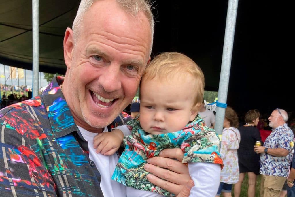A baby in a Hawaiian shirt is looking grumpy as he is being held by the music artist Fat Boy Slim at Camp Bestival in the UK.  It is a family friendly festival.