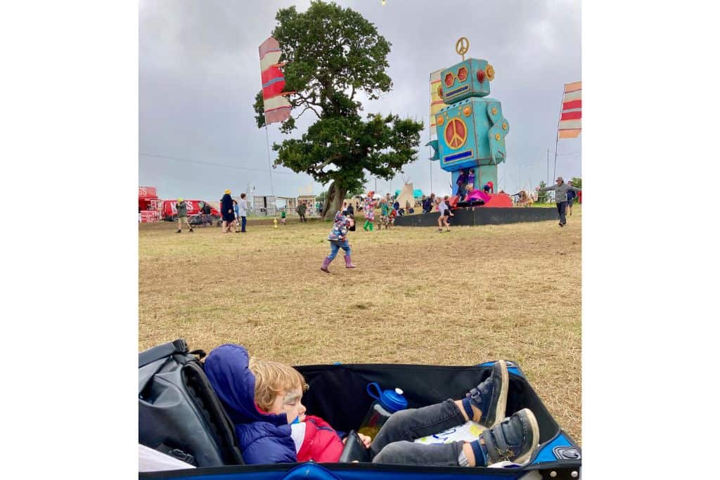 There is a boy lying in a festival trailer side on asleep. In the background is the robot at Camp Bestival in the UK. The boy has been at the family friendly festival all day and fallen asleep. 