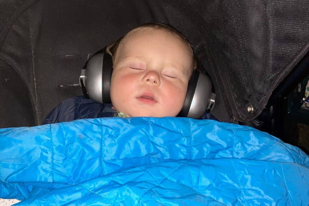 A baby is fast asleep in their pram with ear defenders on and a blue  quilt coat on them as a blanket.