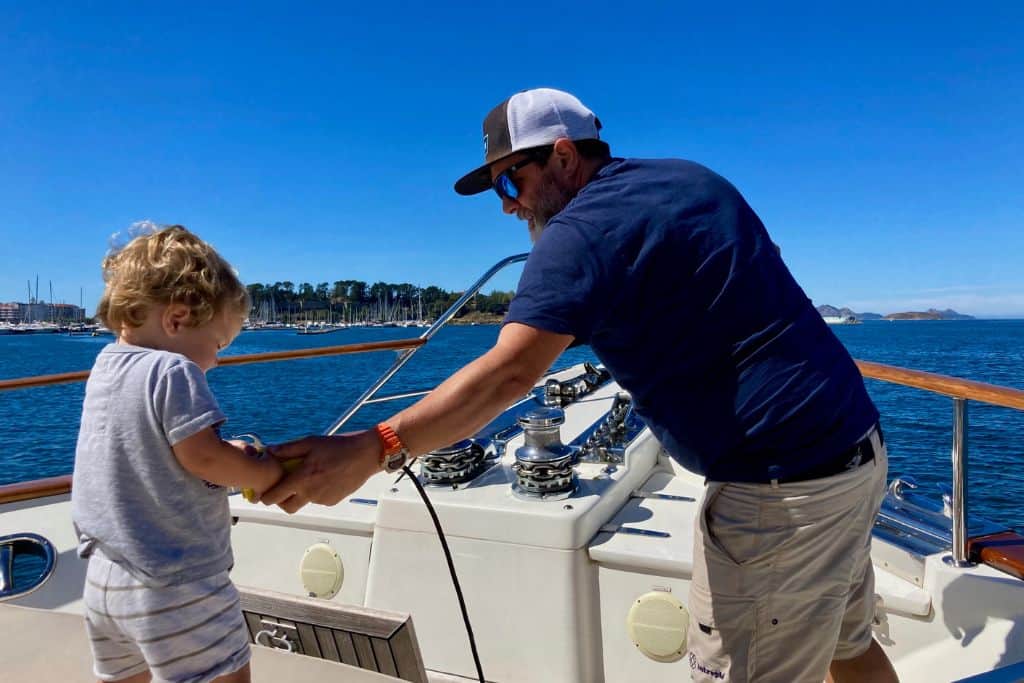 A man is stood at the bow of a yacht with a blue t-shirt on and a baseball cap and sunglasses. Stood next to him in a grey t-shirt is his two year old son who has the electronic keypad for the anchor in his hand. The man is sailing with his kids.