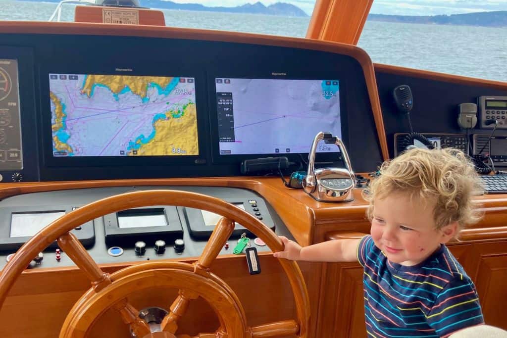 A three year old boy has his hand on the helm of a yacht. He is looking to the side of the shot and not at the camera. By the helm wheel is the navigation computers.  His parents are sailing with their kids.