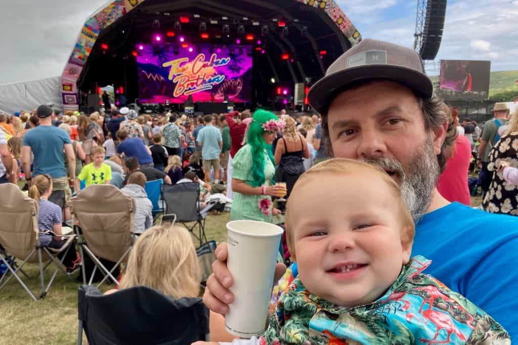 A man is holding a beer and looking at the camera with his baseball cap on. In his arms also looking at the camera is his baby son wearing a Hawaiian shirt.  In the background is the main stage at Camp Bestival and the Cuban Brothers are playing. This is one of the many family friendly festivals in the UK.