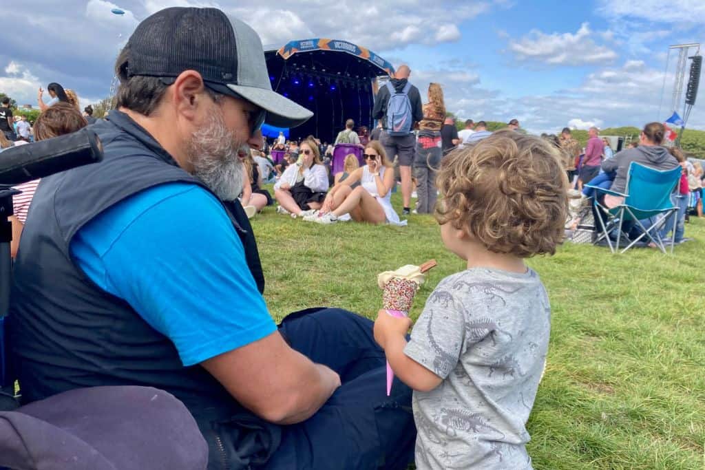 A man in sunglasses and baseball cap is side on and looking at his son who is in a grey t-shirt as he eats an ice cream.  In the background is a music festival stage in the UK.  They are there as it's family friendly.