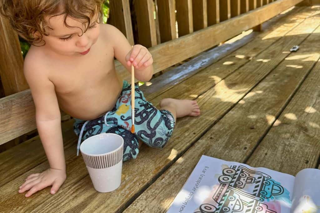 A little boy is sat on some wood decking painting whilst sat in his boardshorts.