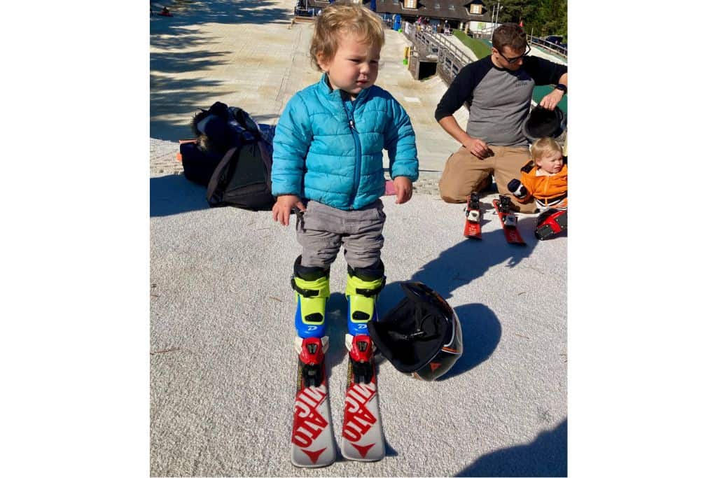 A toddler boy is wearing a green jacket and has skiis on at a dry slope. He is having a skiing lesson with some other kids.