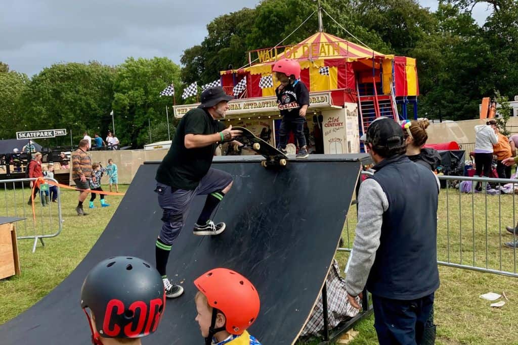 A boy is stood with skate board and helmet at the top of a mini half pipe. There is a man in front of him who is helping him to learn. The boy is just 3 years old and is a Camp Bestival in the UK which is a family friendly festival.