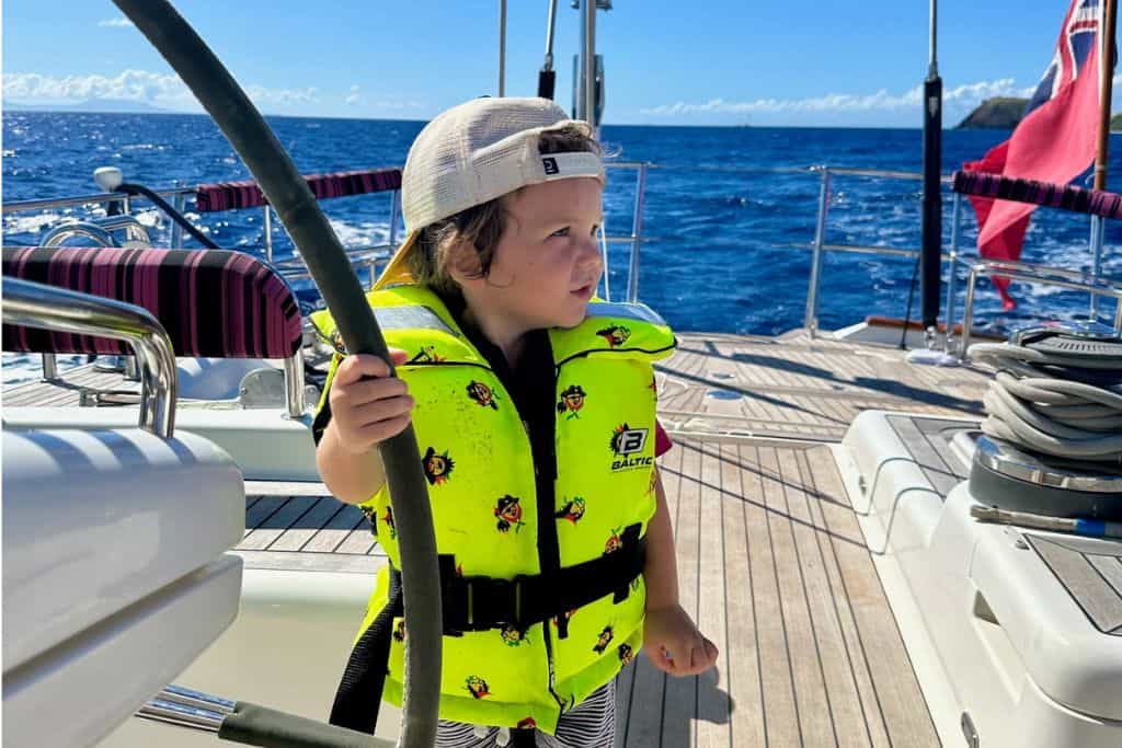 A young boy is wearing a yellow lifejacket with pirates on. His is holding the helm wheel in his hand as he gazes off to the left. He is stood on a sailing yacht.