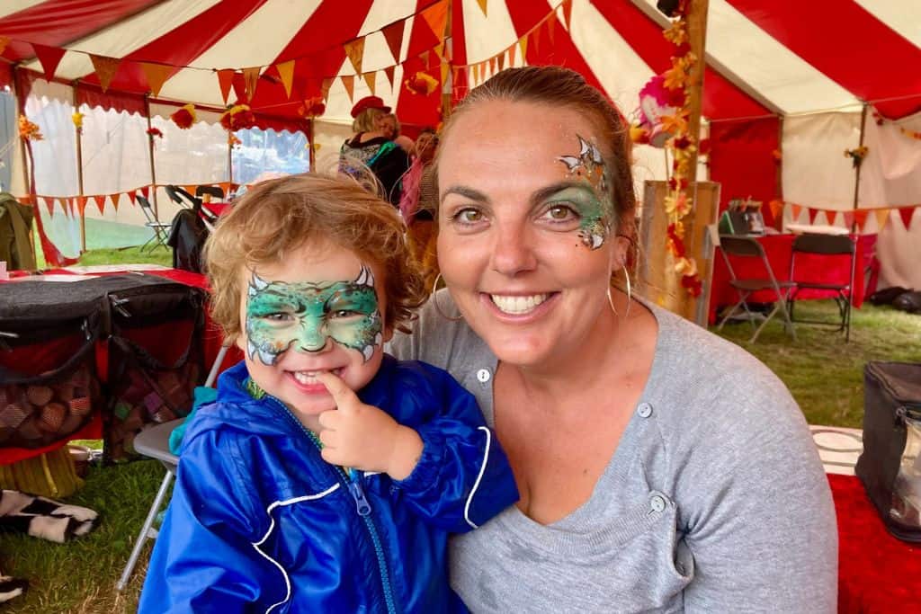 A mum and her son are in a red and white circus tent looking at the camera and smiling.  They both had dinosaur facepaint on.  