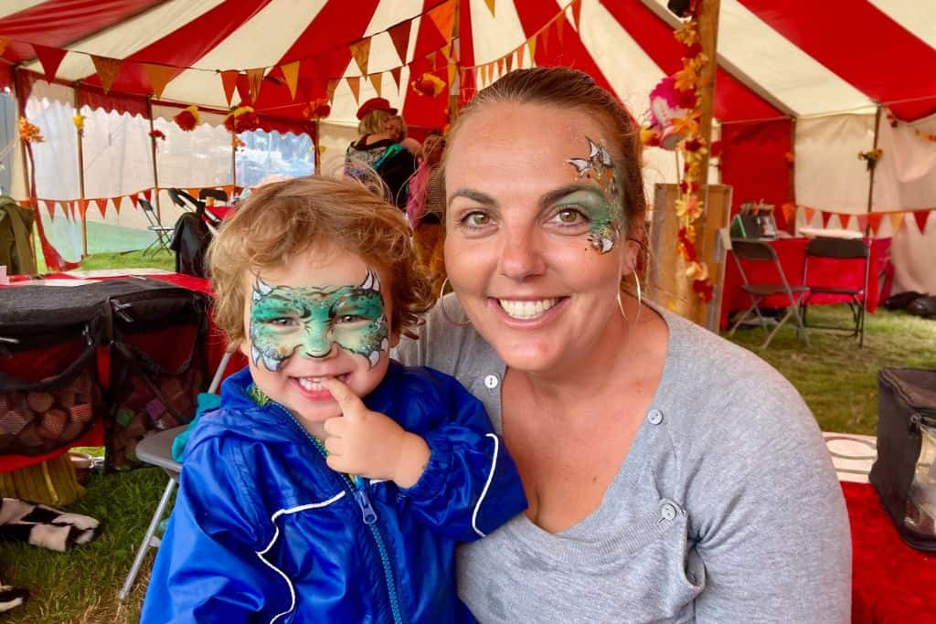 A woman is looking at the camera and has some dragon face paint on. Next to her is her son who also has dragon face paint on. They are in a marquee at Camp Bestival which is one of the best family friendly festivals in the UK.