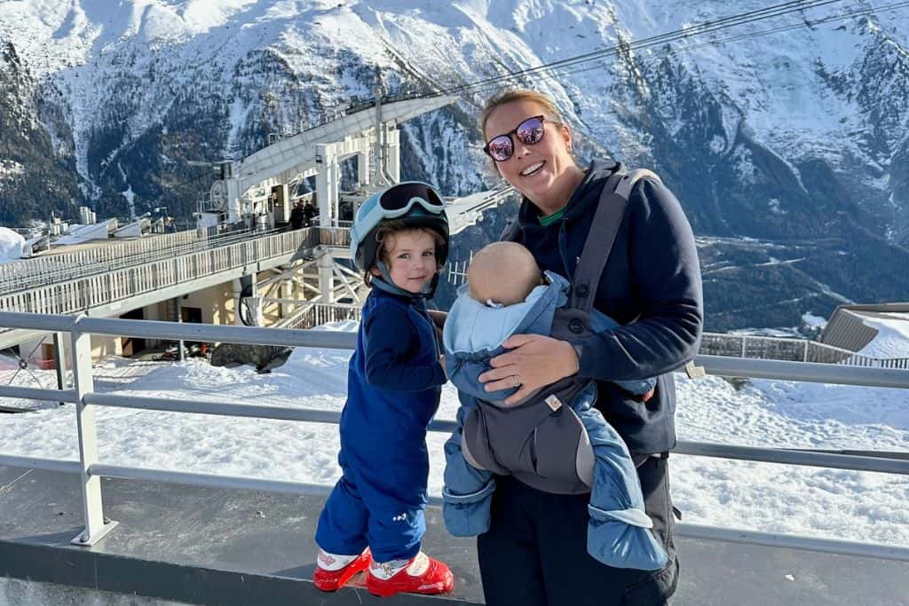 A mum is wearing her baby in a carrier and next to her is her 3 year old son in his ski gear. He is stood on a wall and turning the face the camera with a smile on his face. The mum is skiing with her kids. In the background are the snowcapped mountains and a gondola in the distance.