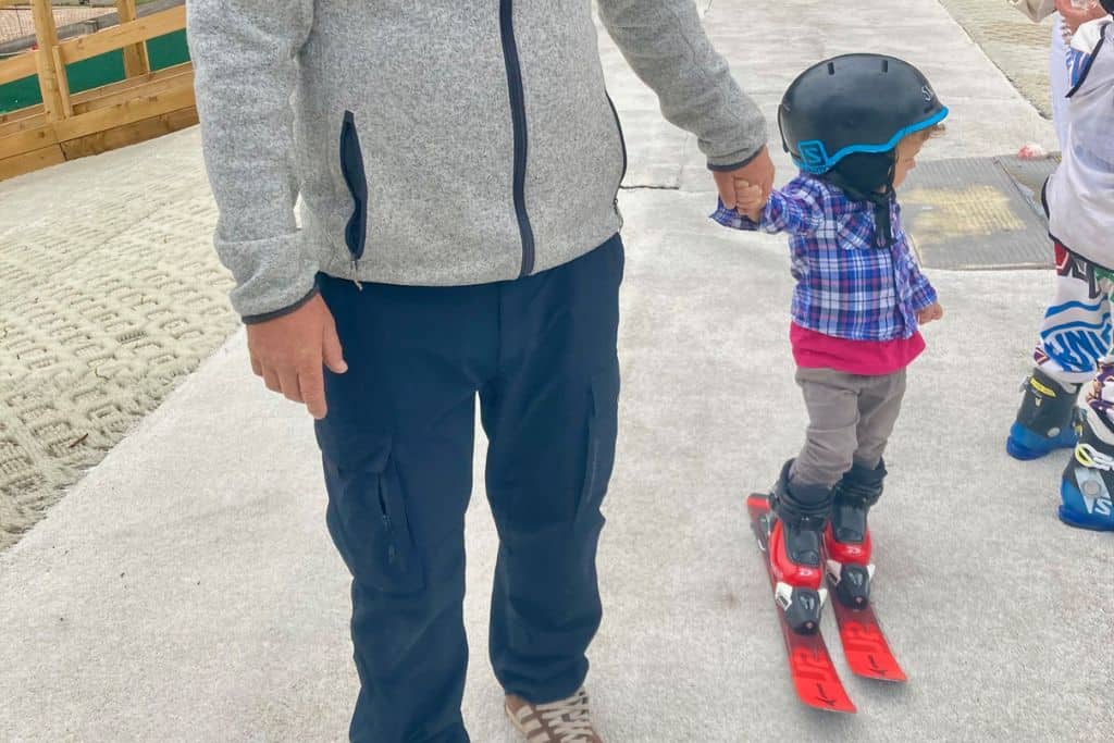 A man is holding the hand of his son who is in ski gear with skis on at the top of a dry ski slope.  The boy is looking off to the side of the photo down the slope. 