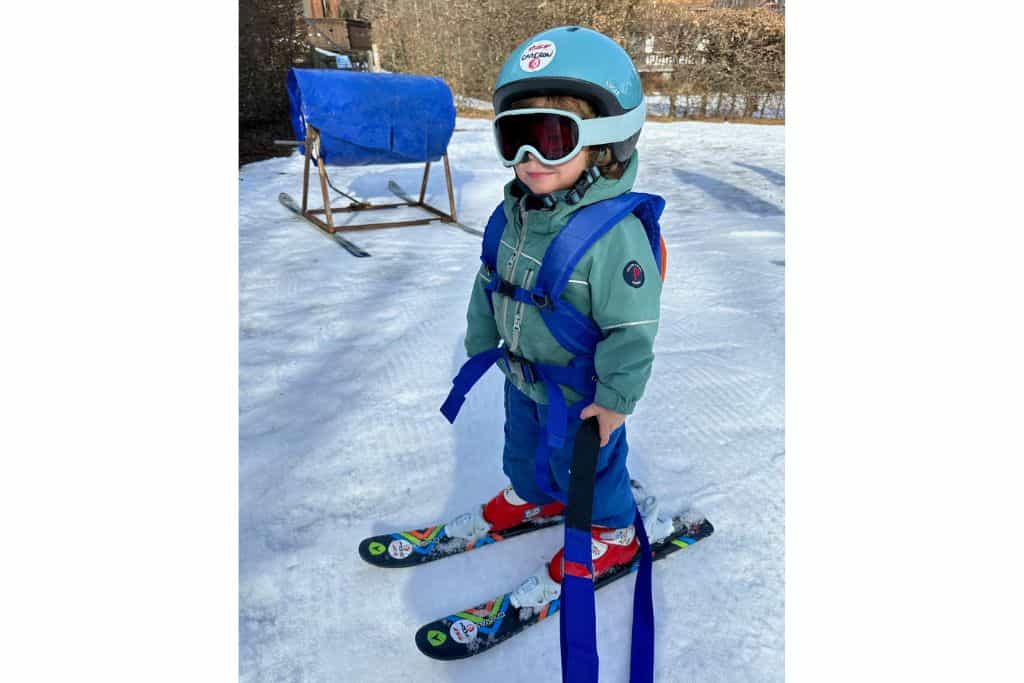 A three year old is standing in his ski gear with his goggles, helmet, and skis's on.  He's also wearing a kids skiing harness about is there with his parents.