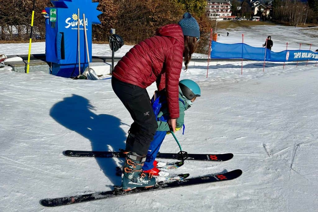 A lady is in her ski clothes and ski's. She has in front of her a toddler boy who also has skis on. She is holding her ski pole out in front of the boy as she's is about to go skiing with him. 