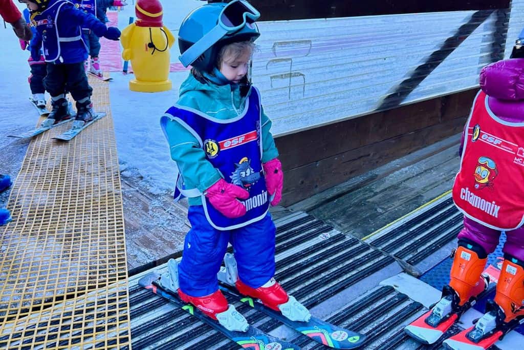 A young boy weating ski gear, skis and a helmet is in a queue in a ski school with other kids and is about to get on the magic carpet to go up the slope.