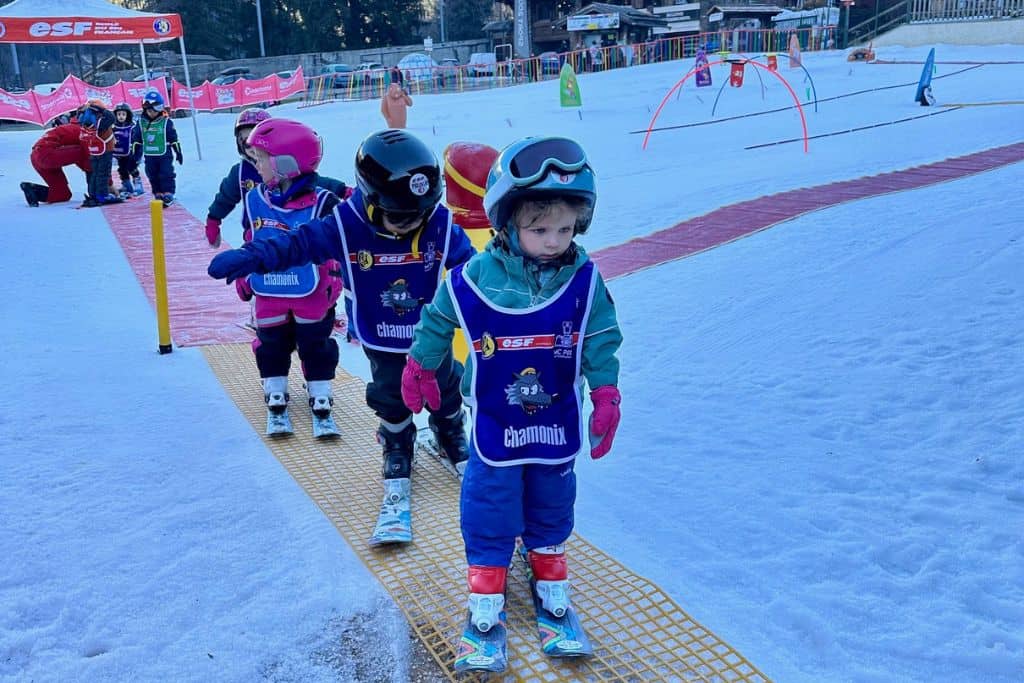 A little toddler boy is at the front of a line of other kids at ski school. He is wearing his ski gear, helmet, goggles and skiis. He is learning to ski with the other kids.