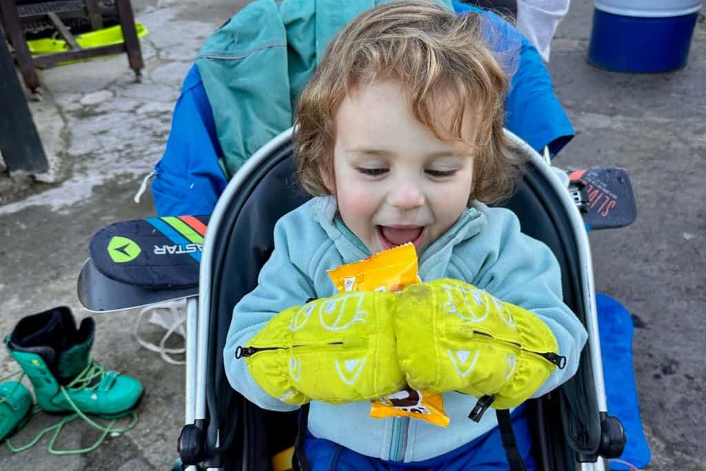 A young boy is sat in his pram in his ski gear and is about to eat a packet of M&M chocolates.  On the back of his pram behind him are his skiis.