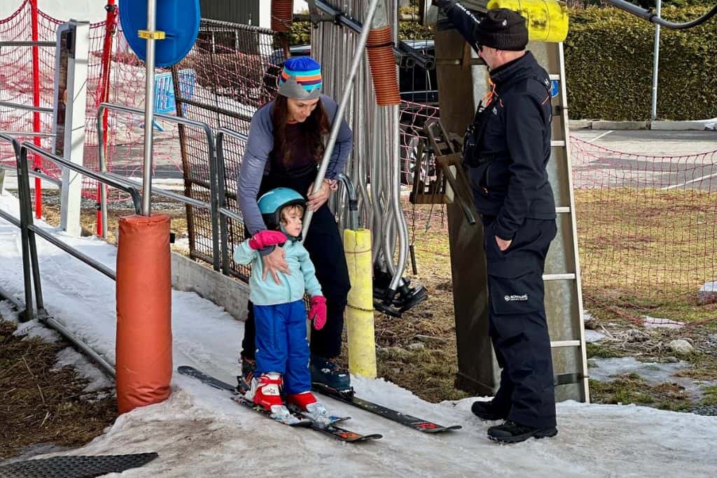 A woman is stood next to a three year old boy and they are both in ski gear with skis on and about to go up a t-bar ski lift.