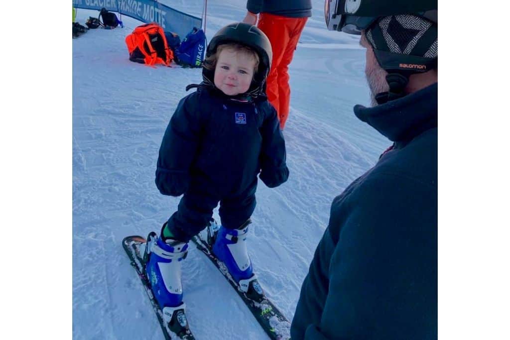 A 2 year old toddler boy has on ski clothes and is stood smiling at the camera with his skiis on and helmet as he is skiing with his parents.  in front of him is his dad.