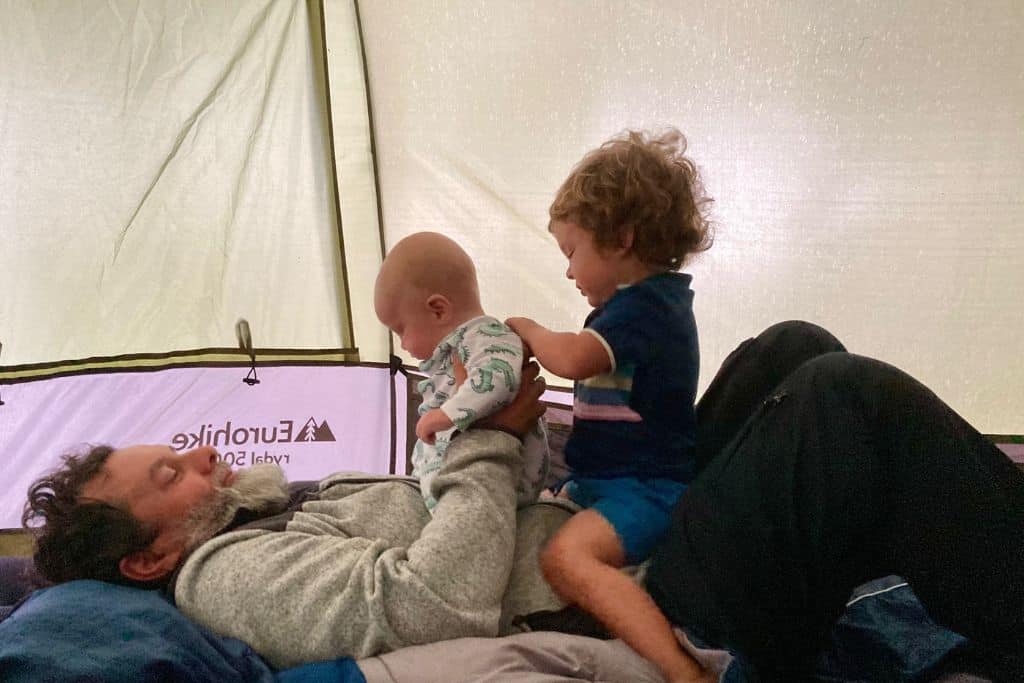 A man is lying on the floor inside a tent and his two son are sat on his tummy laughing.  He is camping with his kids.