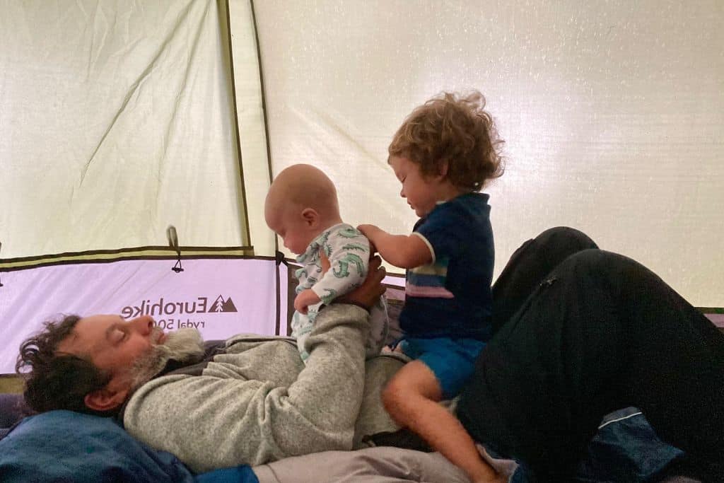 A dad is lying on an airbed in a tent wearing his fleece. Sitting on his tummy are his baby son and toddler son laughing. He is camping with his baby.