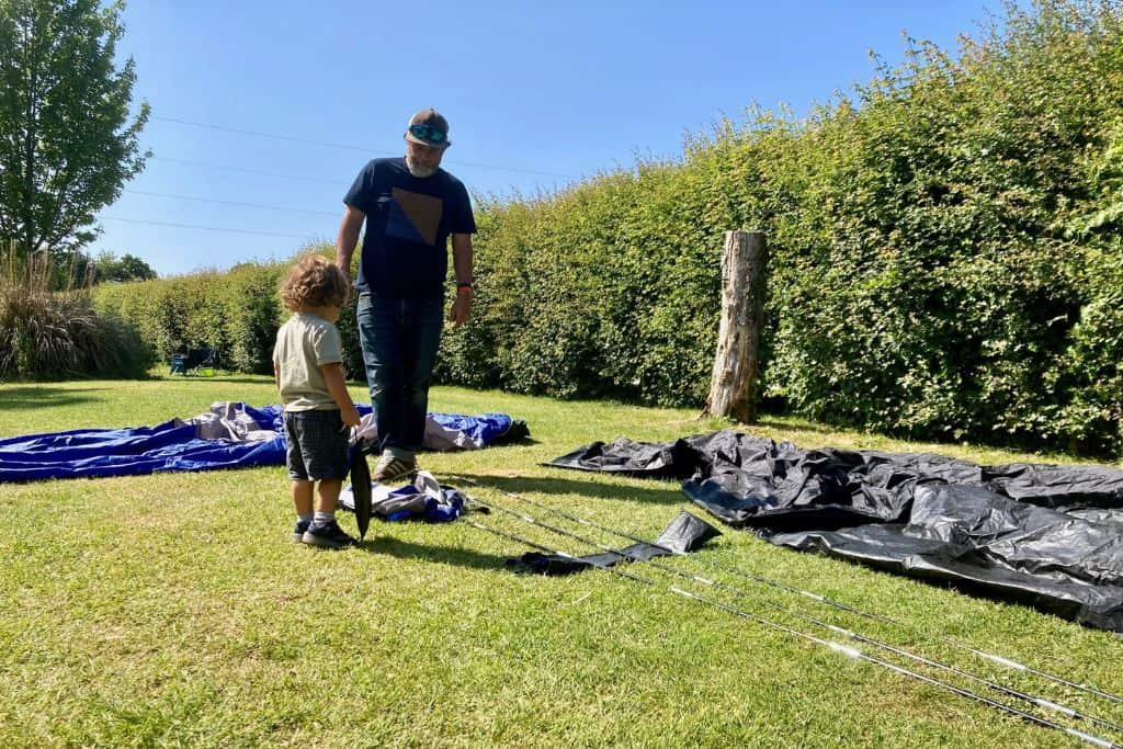 A man and his toddler son are stood in a campsite with the tent laid out on the floor.  They are about to put the tent together. 