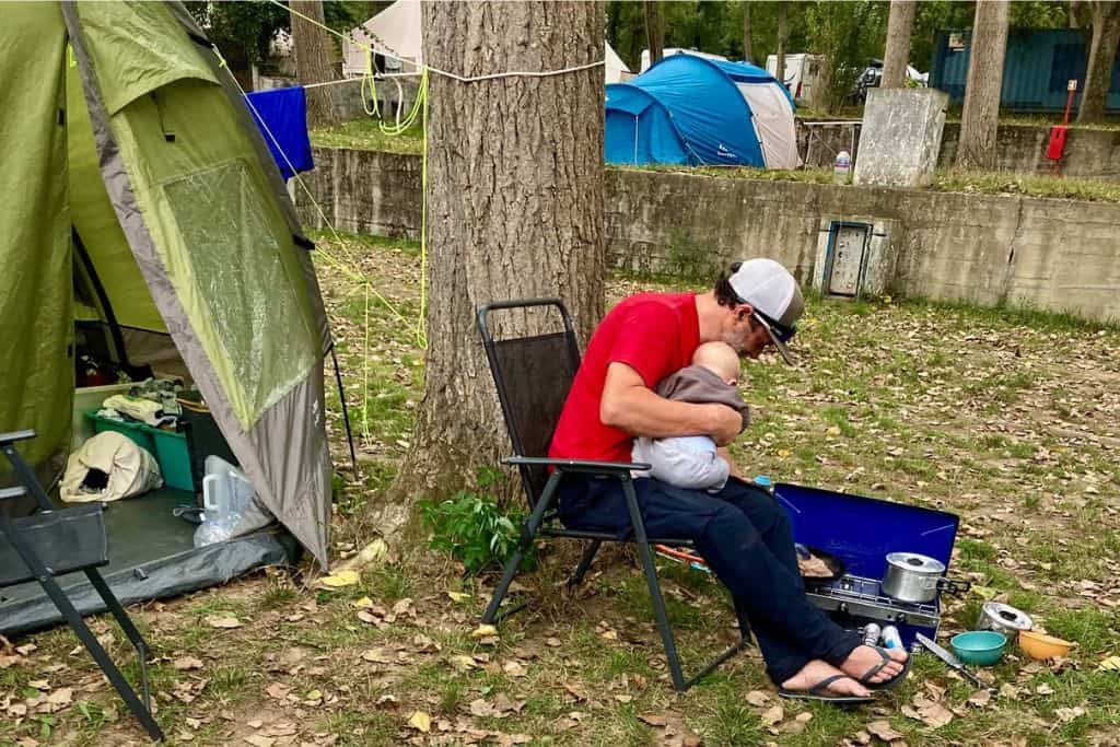 A man is sat in a camping chair with his baby sat on his lap, they have their backs to the viewer and are cooking food on a gas stove. Behind the man is his green tent as he is camping with his kids.