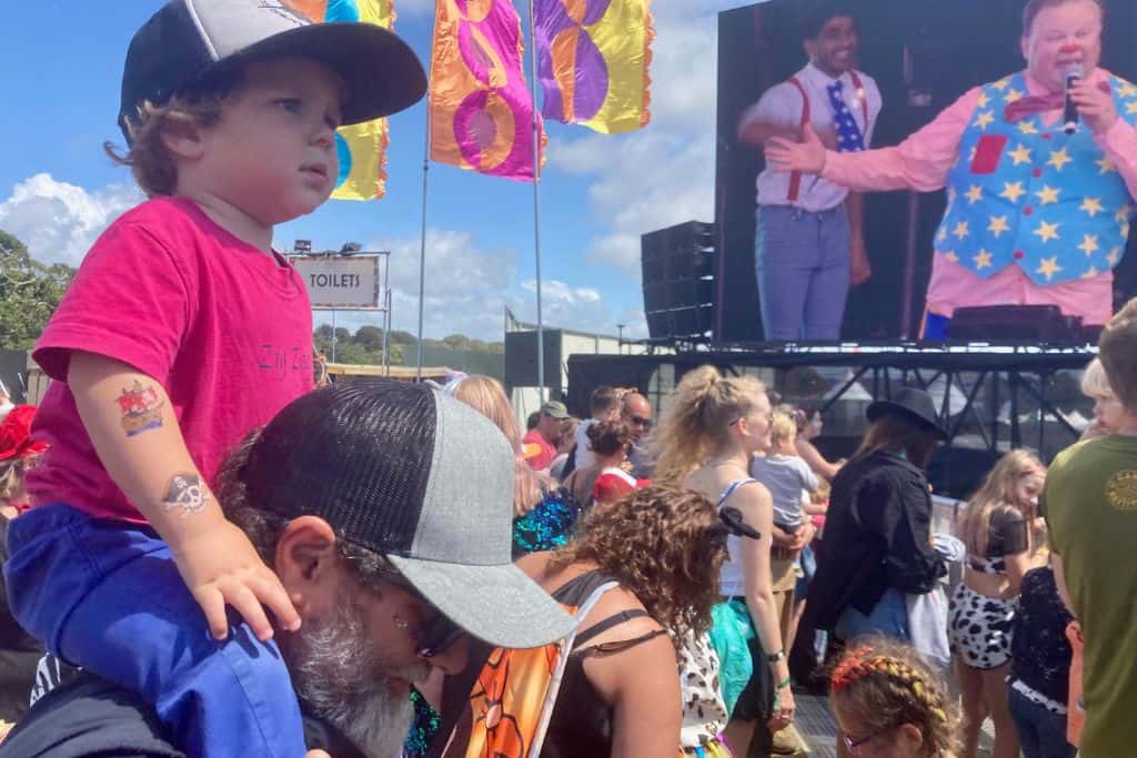 A man in sunglasses and a baseball cap is looking off to the side of the camera. On his shoulders in a pink t-shirt and baseball cap is his son.  They are both looking at the main stage of Camp Bestival in the UK where Mr Tumble is on stage. It is one of the best family friendly festivals,