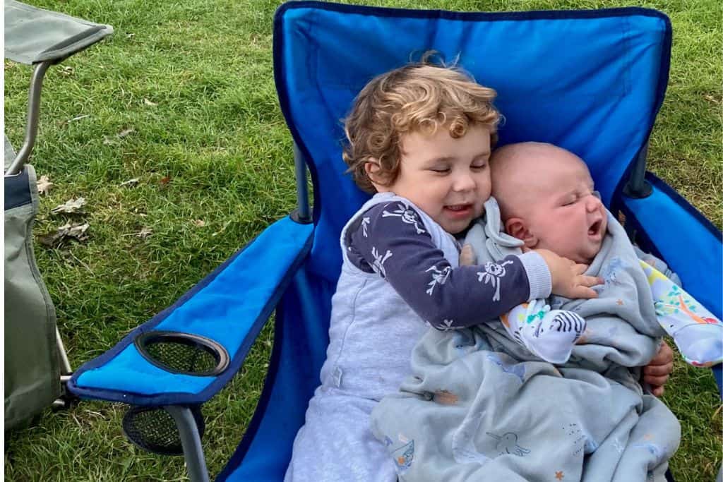 Sat on a bright blue camping chair is a toddler in his sleeping bag and on his lap is his baby brother who is also in a sleeping bag. 