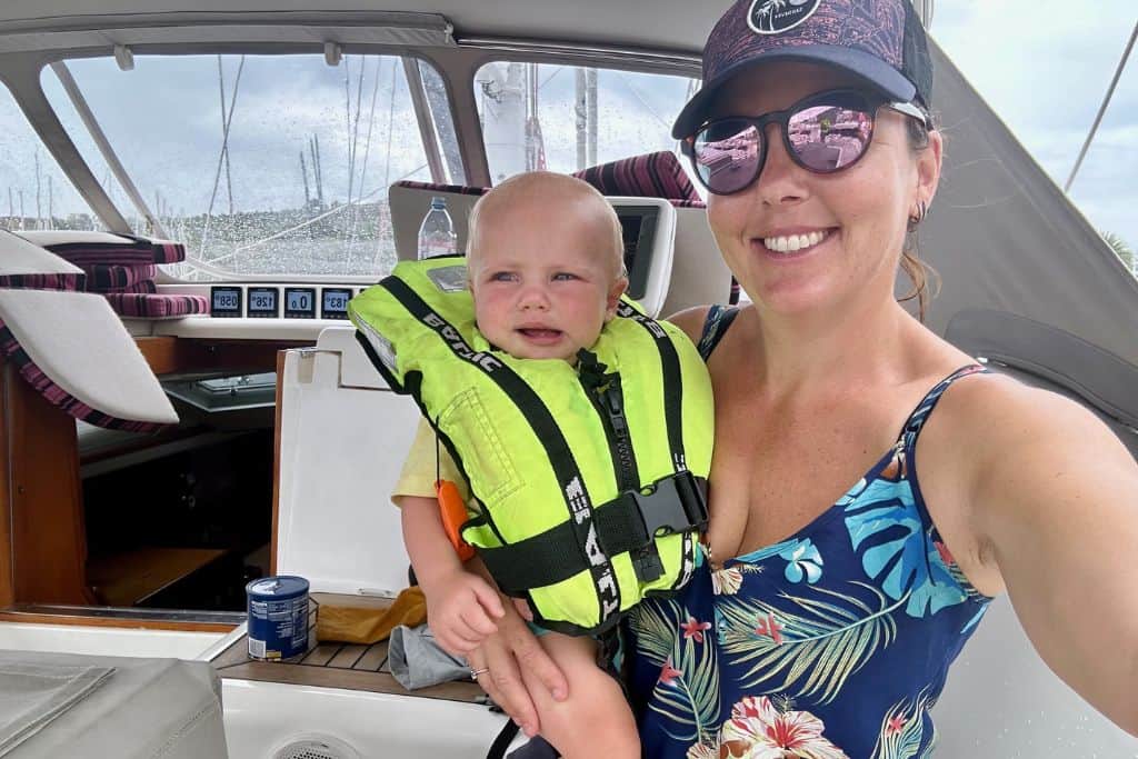 A mum wearing sunglasses and a baseball cap with a flowery vert top is holding her baby son. They are in the cockpit of a yacht and she is sailing with him.