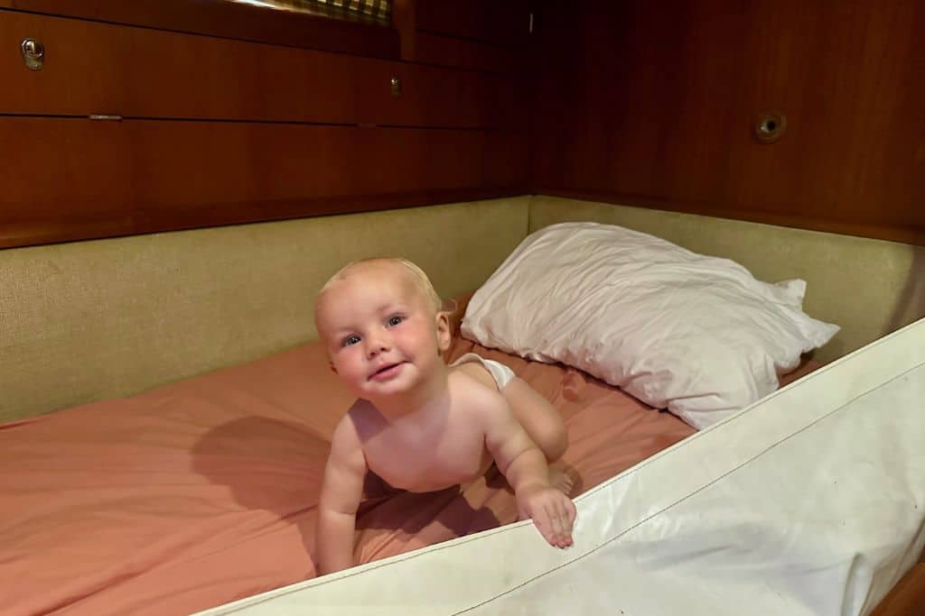 A baby in a nappy is looking at the camera as his playing in a bed on a yacht. His hand is on the top edge of the lee cloth this is hung up so that he can't fall out of the side of the bed. The baby is sailing with his parents.